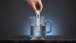 A hand dipping a TDS meter into a glass jug of water on a dark stone ledge.